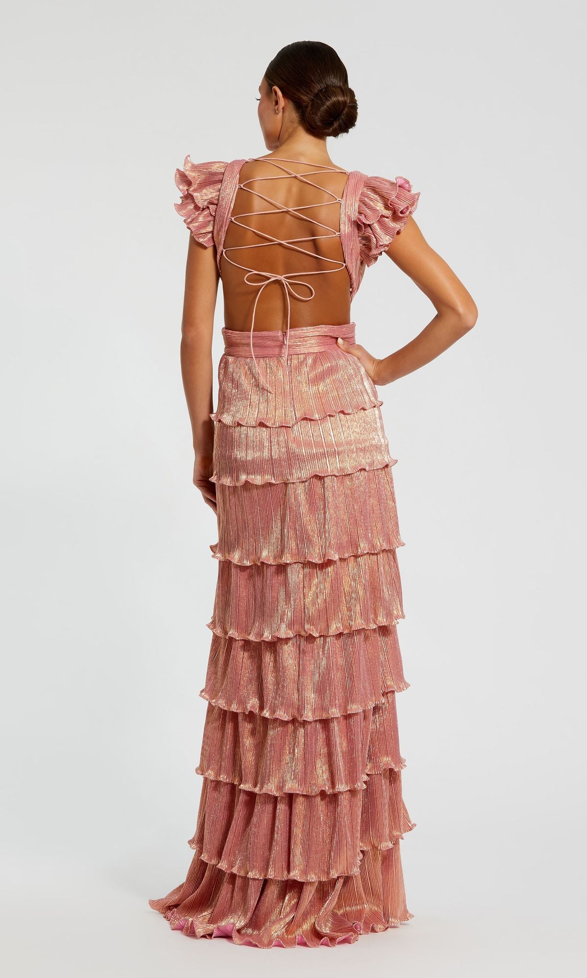 A woman stands with her back to the camera in the Mac Duggal Open-Back Tiered Formal Dress 26740, a pink floor-length gown with flutter sleeves, a lace-up open back, ruffles, and a layered skirt.